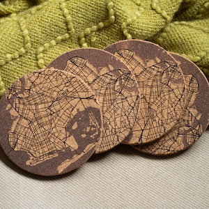 May include: Set of four brown cork coasters featuring a detailed map of a city. The coasters are arranged in a stack on a beige surface.