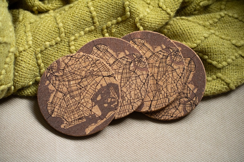 May include: Set of four brown cork coasters featuring a detailed map of a city. The coasters are arranged in a stack on a beige surface.