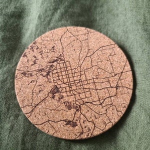 May include: A round cork coaster with a detailed map of a city engraved on it. The map is in black and white and shows the streets and roads of the city.
