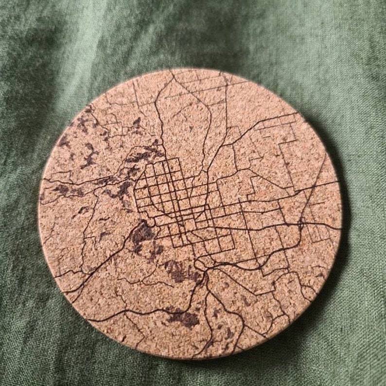 May include: A round cork coaster with a detailed map of a city engraved on it. The map is in black and white and shows the streets and roads of the city.