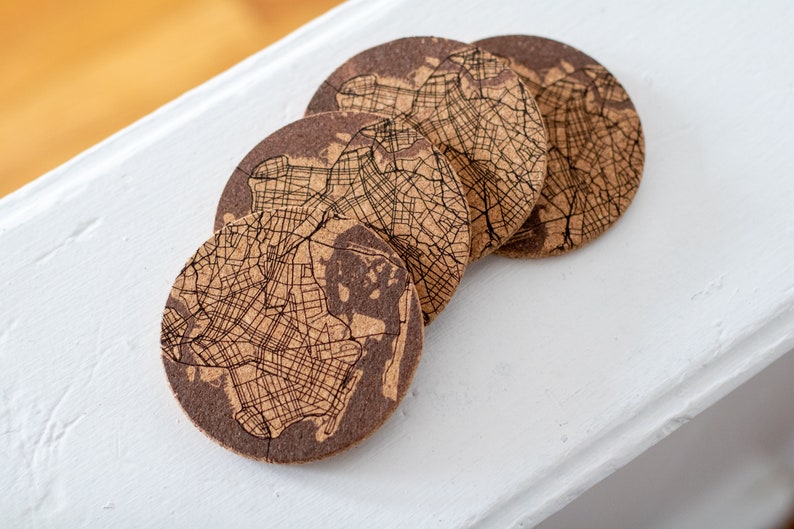 May include: Four cork coasters with a map design. The coasters are brown and feature a detailed map of a city or region.