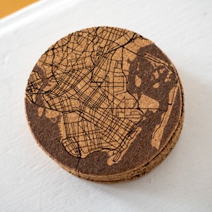 May include: A round cork coaster with a laser-etched map of a city. The coaster is brown and has a natural cork texture.