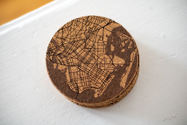 May include: A round cork coaster with a laser-etched map of a city. The coaster is brown and has a natural cork texture.