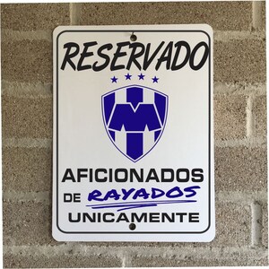 May include: White rectangular sign with black text that reads "RESERVADO" at the top. Below is a blue and white emblem and the text "AFICIONADOS DE RAYADOS UNICAMENTE". The sign is mounted on a brick wall.