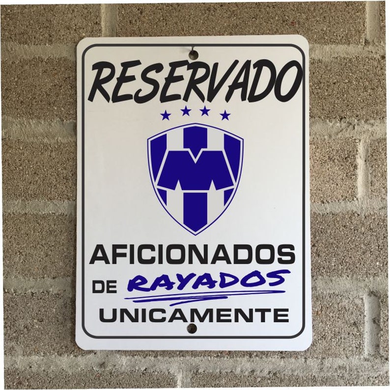 May include: White rectangular sign with black text that reads "RESERVADO" at the top. Below is a blue and white emblem and the text "AFICIONADOS DE RAYADOS UNICAMENTE". The sign is mounted on a brick wall.