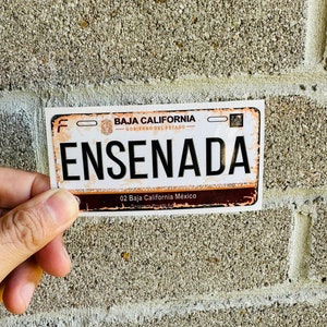 May include: A white and brown license plate sticker with the text "BAJA CALIFORNIA" and "GOBIERNO DEL ESTADO" in orange. The sticker also has the text "ENSENADA" in black letters. The sticker has a brown border and the text "02 Baja California México" in white letters.