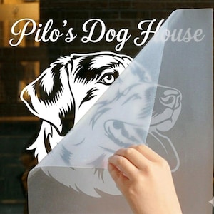 May include: A white vinyl decal featuring a detailed illustration of a dog's head, partially covered by a transparent backing. The text "Pilo's Dog House" is arched above the dog illustration in a cursive font.