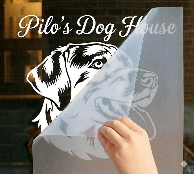 May include: A white vinyl decal featuring a detailed illustration of a dog's head, partially covered by a transparent backing. The text "Pilo's Dog House" is arched above the dog illustration in a cursive font.