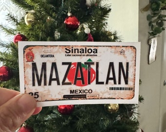 May include: A white and red license plate sticker with the text "Sinaloa Lider nacional en alimentos" and "Mazatlan" in black letters. The sticker also includes the text "Mexico" and "Delantera" in smaller font.