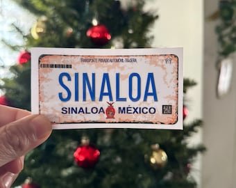 May include: A rectangular sticker resembling a license plate, featuring the word "SINALOA" in large blue letters. Below, it reads "SINALOA MÉXICO." The background has a distressed, vintage look with a faded brown and white color scheme. A Christmas tree with red ornaments is in the background.