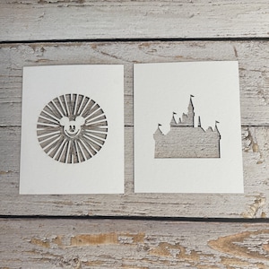May include: Two white paper cutouts on a wooden surface. One features a Mickey Mouse face surrounded by sun rays. The other is a silhouette of a castle with flags. The cutouts are likely for crafting or decoration.