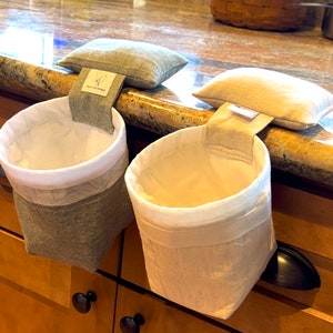 Op de afbeelding: Twee stoffen hangmanden met witte binnenkant en crèmekleurige buitenkant worden getoond. Elke mand heeft een stoffen lus en een klein kussen bovenop. De manden hangen aan een aanrecht. De manden zijn ontworpen om aan een kastdeur te hangen.