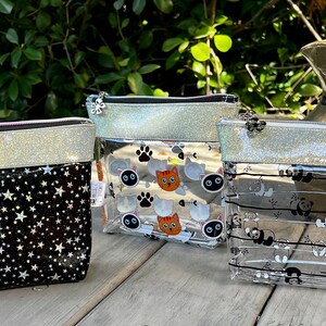 Könnte beinhalten: Drei transparente Reißverschlusstaschen mit silbernem Glitzerbesatz. Die Taschen haben verschiedene Muster: schwarze Sterne, Katzen und Pfotenabdrücke sowie Pandas.