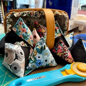 May include: A collection of handmade fabric weights in various floral patterns and solid black, arranged near a wooden basket. A blue OLFA rotary cutter with a yellow blade is in the foreground, suggesting a sewing or quilting project.