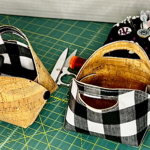 May include: Two small fabric baskets with black and white checkered fabric and cork bottoms. The baskets have handles and are sitting on a green cutting mat.