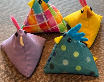 May include: Four colorful fabric chick toys. The chicks are triangular shaped and have black eyes. One chick is purple, one is yellow, one is teal with white polka dots, and one is multi-colored with a checkered pattern.