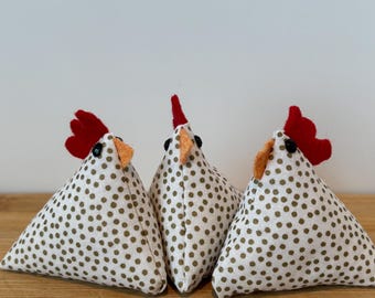 May include: Three white triangular fabric chickens with brown polka dots, red combs, and orange beaks. The chickens are sitting on a wooden surface.