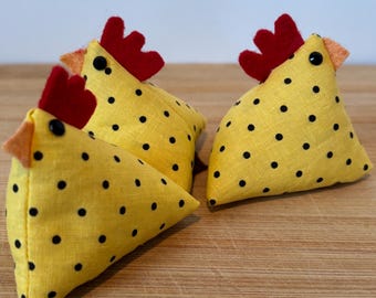 May include: Three yellow fabric chickens with black polka dots and red combs. The chickens are sitting on a wooden surface.