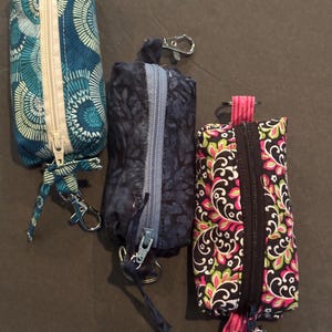 May include: Three fabric pouches with zippers and keychains. The pouches are cylindrical and feature different patterns: teal and white swirls, navy blue with a leaf design, and black with pink and green floral patterns.