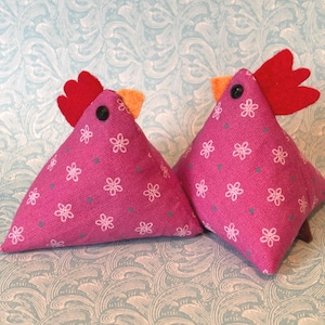 May include: Two pink fabric chicken-shaped decorations. Each has a red felt comb, an orange wattle, and a black button eye. The chickens are decorated with white floral patterns and small blue dots.