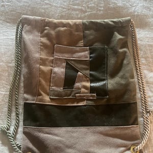 May include: A drawstring backpack with a patchwork design in shades of brown, tan, and olive green. The bag features a small square pocket and a dark green band across the middle. The drawstring is a light beige rope.