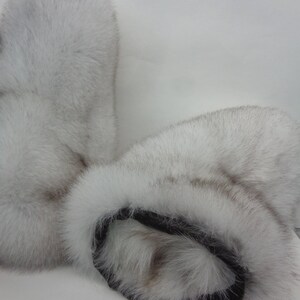 May include: A pair of white fur mittens with a black leather cuff. The mittens are made of soft, fluffy fur and appear to be warm and cozy. The fur has a slight greyish hue.