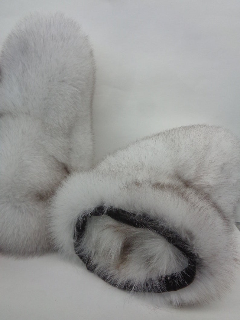 May include: A pair of white fur mittens with a black leather cuff. The mittens are made of soft, fluffy fur and appear to be warm and cozy. The fur has a slight greyish hue.