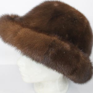 May include: A brown fur hat with a rounded crown and a wide, slightly upturned brim. The hat is made of soft, plush fur and is displayed on a white mannequin head. The fur appears to be a rich, dark brown color.