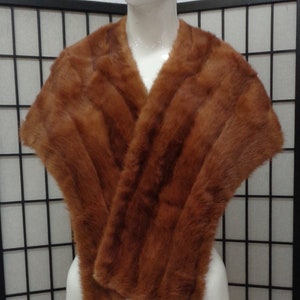 May include: A brown fur shawl with a soft, luxurious texture. The shawl is draped over a white mannequin and is folded in a way that shows the different layers of fur.