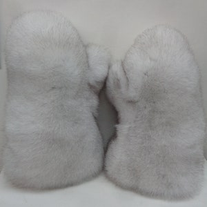 May include: A pair of light gray fur mittens. The mittens are made of soft, fluffy fur and have a rounded shape. The fur appears to be a natural color, with variations in tone. The mittens are designed to keep hands warm in cold weather.