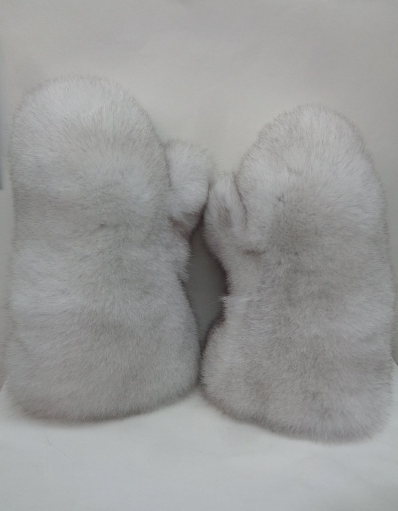 May include: A pair of light gray fur mittens. The mittens are made of soft, fluffy fur and have a rounded shape. The fur appears to be a natural color, with variations in tone. The mittens are designed to keep hands warm in cold weather.