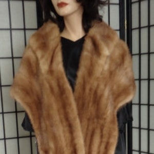 May include: A brown fur shawl with a wide collar. The shawl is draped over a black dress on a mannequin. The fur appears soft and has a gradient of light and dark brown tones.