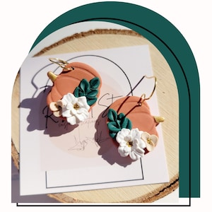 May include: A pair of pumpkin-shaped earrings with white flowers and green leaves. The earrings are made of polymer clay and have gold hooks.