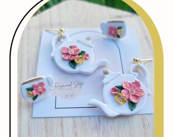 May include: A set of three white ceramic earrings shaped like teapots and teacups, decorated with pink and yellow flowers and gold accents. The earrings are displayed on a white card with the text "Rosewood Stage".