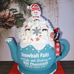 May include: A blue teapot with white snow-like frosting and a snowman on top. The teapot is decorated with the words "Snowball Falls - Cafe and Bakery - Hot Chocolate Served Fresh Daily".