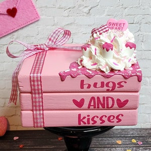 Puede incluir: Caja de madera rosa con un diseño de glaseado blanco y rosa. La caja está apilada sobre un pedestal negro y tiene las palabras "abrazos y besos" escritas en ella. La caja está rodeada de corazones rosas.