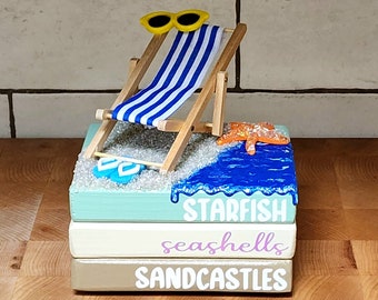 May include: A stack of three wooden blocks with a beach theme. The top block has a blue, white, and yellow beach chair with sunglasses, a starfish, and blue water. The middle block says "seashells" in purple lettering. The bottom block says "sandcastles" in white lettering.