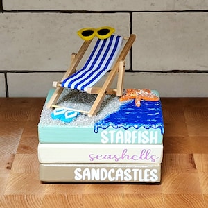 May include: A stack of three wooden blocks with a beach theme. The top block has a blue, white, and yellow beach chair with sunglasses, a starfish, and blue water. The middle block says "seashells" in purple lettering. The bottom block says "sandcastles" in white lettering.