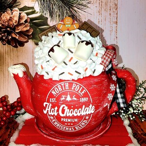May include: Red ceramic teapot with a white and black checkered ribbon, filled with white marshmallows, chocolate sauce, and sprinkles. The teapot has a white label that reads "North Pole Est. 1897 Hot Chocolate Premium Christmas Blend".