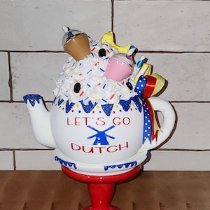 May include: White teapot with blue and red trim, decorated with white frosting, sprinkles, and a windmill. The teapot is on a red pedestal and says "Let's Go Dutch".