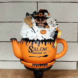 May include: Orange teapot with black dripping paint and a black stand. The teapot has a black and white owl with a witch hat on top. The teapot has the text "Salem Broom Co." on the side.