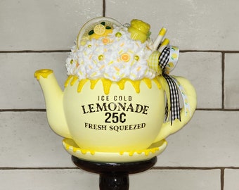 May include: A yellow teapot with a white lid and a black base. The teapot is decorated with lemons, flowers, and a black and white checkered ribbon. The teapot has a sign that reads "ICE COLD LEMONADE 25¢ FRESH SQUEEZED".