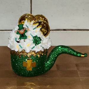 May include: A green and gold glitter pipe with a white and green shamrock design. The pipe is filled with white frosting and topped with a small leprechaun figurine and gold coins.