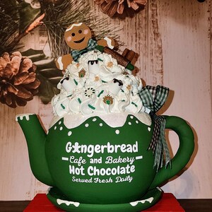 May include: Green ceramic teapot with a gingerbread man and whipped cream on top. The teapot has a white inscription that reads "Gingerbread - Cafe and Bakery - Hot Chocolate - Served Fresh Daily".