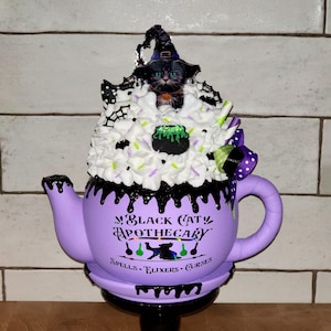 May include: A whimsical, purple apothecary teapot with a black base, featuring a black cat wearing a witch hat atop a whipped cream design. The teapot is decorated with the text "Black Cat Apothecary" and the words "Spells, Elixirs, Curses."