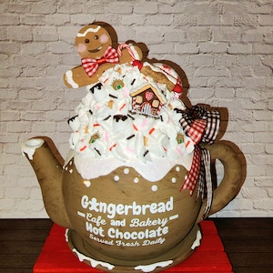 May include: A brown ceramic teapot decorated with white frosting, sprinkles, and a gingerbread house. A gingerbread man with a red bow tie sits on top of the teapot. The teapot has a white sign that reads "Gingerbread Cafe and Bakery - Hot Chocolate Served Fresh Daily".
