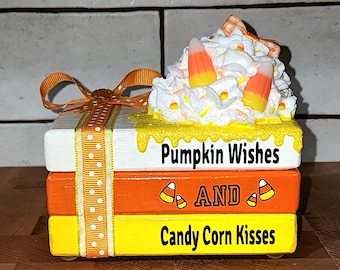 Puede incluir: Una pila de tres bloques de madera decorados con diseños de otoño. El bloque superior es blanco con pintura amarilla que gotea y el texto "Pumpkin Wishes". El bloque del medio es naranja con el texto "AND" y dos imágenes de maíz dulce. El bloque inferior es amarillo con el texto "Candy Corn Kisses". Los bloques están unidos con una cinta blanca con lunares naranjas.