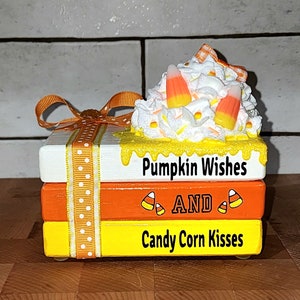 Puede incluir: Una pila de tres bloques de madera decorados con diseños de otoño. El bloque superior es blanco con pintura amarilla que gotea y el texto "Pumpkin Wishes". El bloque del medio es naranja con el texto "AND" y dos imágenes de maíz dulce. El bloque inferior es amarillo con el texto "Candy Corn Kisses". Los bloques están unidos con una cinta blanca con lunares naranjas.