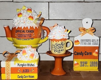 Puede incluir: Un conjunto de decoraciones de otoño con temática de maíz dulce que incluye una tetera de maíz dulce, una taza de maíz dulce, una pila de libros con detalles de maíz dulce y un letrero de madera que dice "Sweeter Than Candy Corn Est. 1898".
