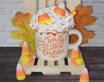 Puede incluir: Taza de cerámica blanca con un diseño de maíz dulce y el texto "Candy Corn Cutie". La taza está llena de crema batida y maíz dulce. La taza está sentada en una caja de madera con hojas de otoño y maíz dulce esparcidos a su alrededor.